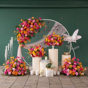 Könnte beinhalten: Eine lebendige Blumendekoration mit rosa, orangefarbenen und gelben Rosen, angeordnet auf weißen Sockeln und einem dekorativen Bogen. Die Präsentation umfasst Kerzen, eine Papiertüte mit Grün und eine große Schmetterlingsdekoration.