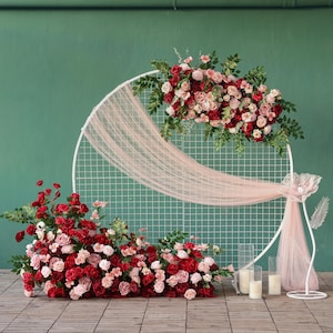 Könnte beinhalten: Ein weißer Hochzeitsbogen aus Metall mit Gittermuster, verziert mit roten und rosa Rosen und Grün. Ein durchsichtiger rosa Stoff drapiert sich über den Bogen. Zusätzliche Blumengestecke und Kerzen befinden sich an der Basis.