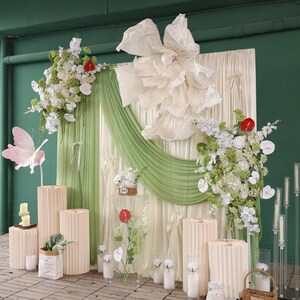 May include: A decorative backdrop for an event, featuring a large paper flower, green draping, and floral arrangements. The scene includes beige columns, a paper butterfly, and candles, creating an elegant and natural aesthetic.