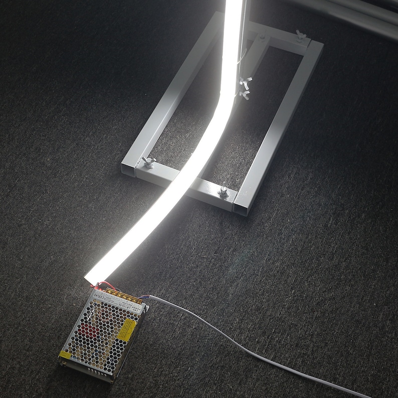 K&ouml;nnte beinhalten: Ein wei&szlig;er LED-Lichtstreifen ist zu einer gebogenen Form gebogen und auf einem wei&szlig;en Metallst&auml;nder montiert. Ein wei&szlig;es Stromkabel verbindet den Lichtstreifen mit einem schwarzen Netzteil.