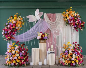 Pode incluir: Uma composição floral decorativa com uma grande borboleta, tecido drapeado e vários arranjos florais. As flores são de várias cores, incluindo rosa, amarelo e vermelho. O fundo inclui cortinas brancas e rosa e pilares decorativos.
