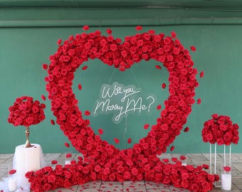 May include: A heart-shaped floral arch made of red roses with the words "Will you marry me?" written on a chalkboard behind it. The arch is decorated with red rose petals on the floor.