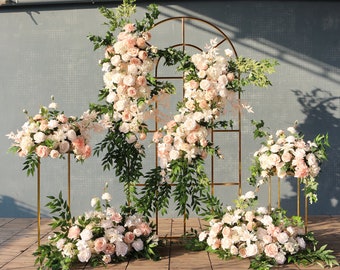May include: A wedding backdrop with a gold archway decorated with white and blush pink flowers and greenery. The archway is flanked by two smaller arrangements of flowers on gold stands.