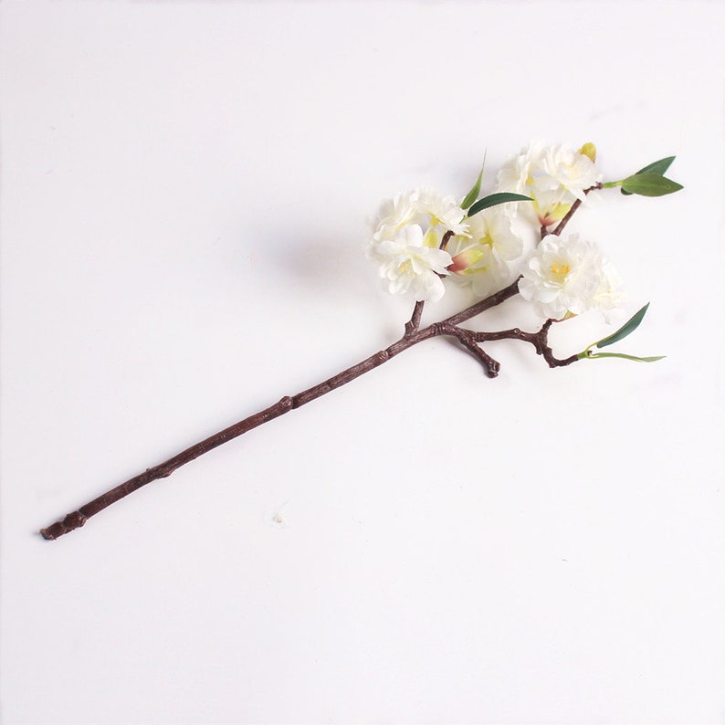 May include: A single white artificial flower branch with multiple blossoms and green leaves. The branch is brown and has a natural, rustic look.