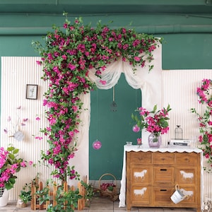 Puede incluir: Una exhibición decorativa con vibrantes flores rosas y follaje verde que caen sobre un arco. Un mueble de madera con diseños de caballos y un jarrón de flores también están presentes. La escena se desarrolla sobre un fondo verde y blanco.
