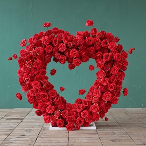 Könnte beinhalten: Ein großes herzförmiges Blumenarrangement aus zahlreichen roten Rosen. Die Rosen sind dicht gepackt und bilden die Form eines Herzens mit einer offenen Mitte. Das Arrangement steht auf einer weißen Basis vor einer türkisblauen Wand und einem Holzboden.