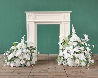 May include: A white decorative fireplace mantel against a green backdrop. Two large displays of white flowers and foliage are positioned on the floor in front of the mantel. The floor is made of wooden planks.