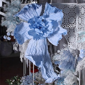 May include: A large, light blue paper flower with ruffled petals. The flower is hanging from a white metal frame.