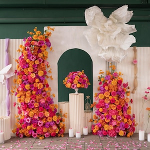 May include: A vibrant floral arrangement featuring a mix of orange, pink, and fuchsia flowers. The display includes two large floral structures, a smaller arrangement on a pedestal, and decorative elements, creating a colorful and festive scene.