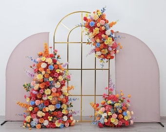 May include: A gold metal archway decorated with colorful artificial flower arrangements. The archway is flanked by two pink fabric panels. The flowers are a mix of orange, yellow, pink, red, and blue.