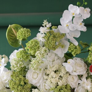 May include: A close-up of a floral arrangement featuring white roses, orchids, and green accents. The arrangement includes various textures and shades of green, with a backdrop of a dark green surface.