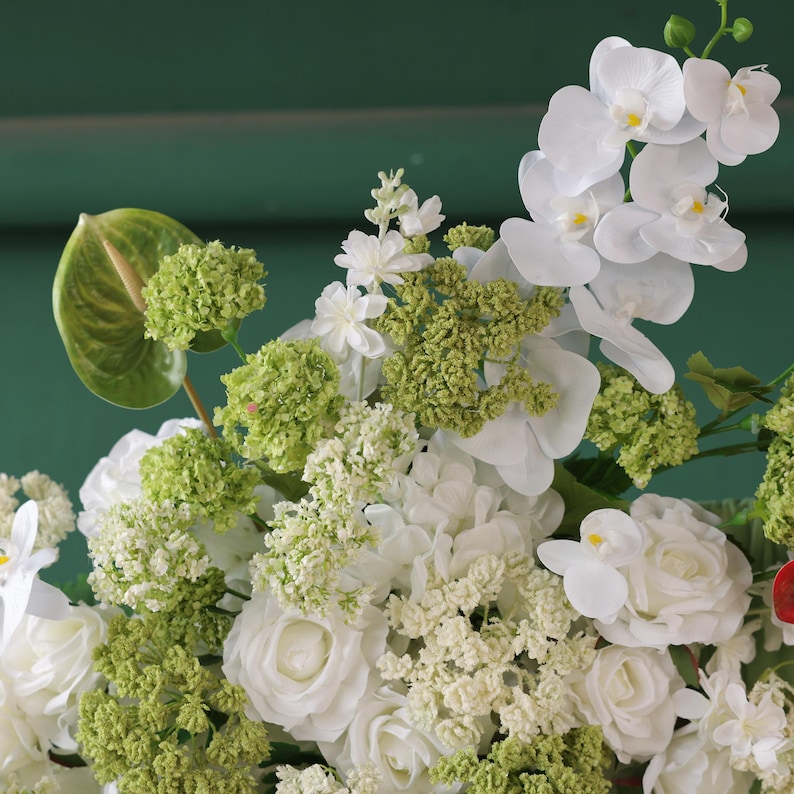 May include: A close-up of a floral arrangement featuring white roses, orchids, and green accents. The arrangement includes various textures and shades of green, with a backdrop of a dark green surface.