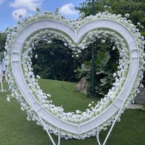 May include: White heart-shaped floral archway made of metal with white flowers and greenery. The archway is standing on a green grassy lawn.