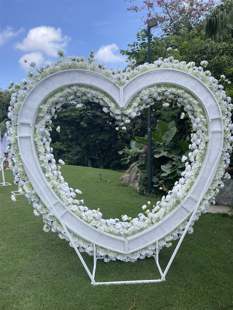 May include: White heart-shaped floral archway made of metal with white flowers and greenery. The archway is standing on a green grassy lawn.