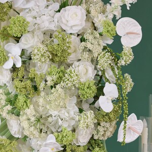 May include: A lush floral arrangement featuring white roses, orchids, and anthuriums, accented with vibrant green foliage. The composition is dense and full, with a variety of textures and shapes, creating a visually rich display. The background is a solid, deep green.