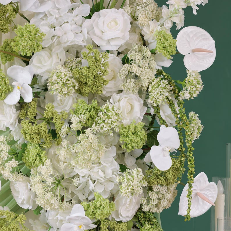 May include: A lush floral arrangement featuring white roses, orchids, and anthuriums, accented with vibrant green foliage. The composition is dense and full, with a variety of textures and shapes, creating a visually rich display. The background is a solid, deep green.