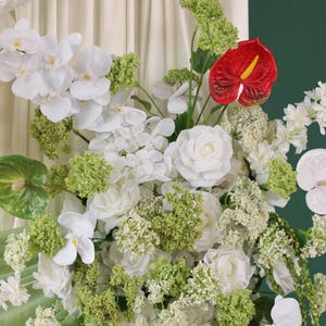 May include: A lush floral arrangement featuring white orchids, roses, and other white flowers, accented with green foliage and a vibrant red anthurium. The flowers are arranged against a soft, neutral background.