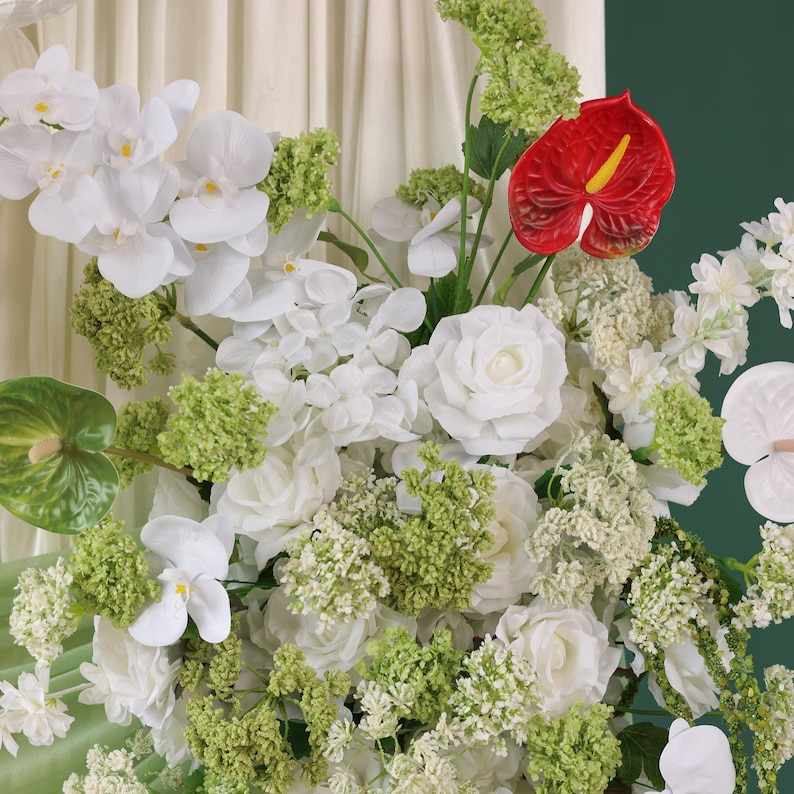 May include: A lush floral arrangement featuring white orchids, roses, and other white flowers, accented with green foliage and a vibrant red anthurium. The flowers are arranged against a soft, neutral background.