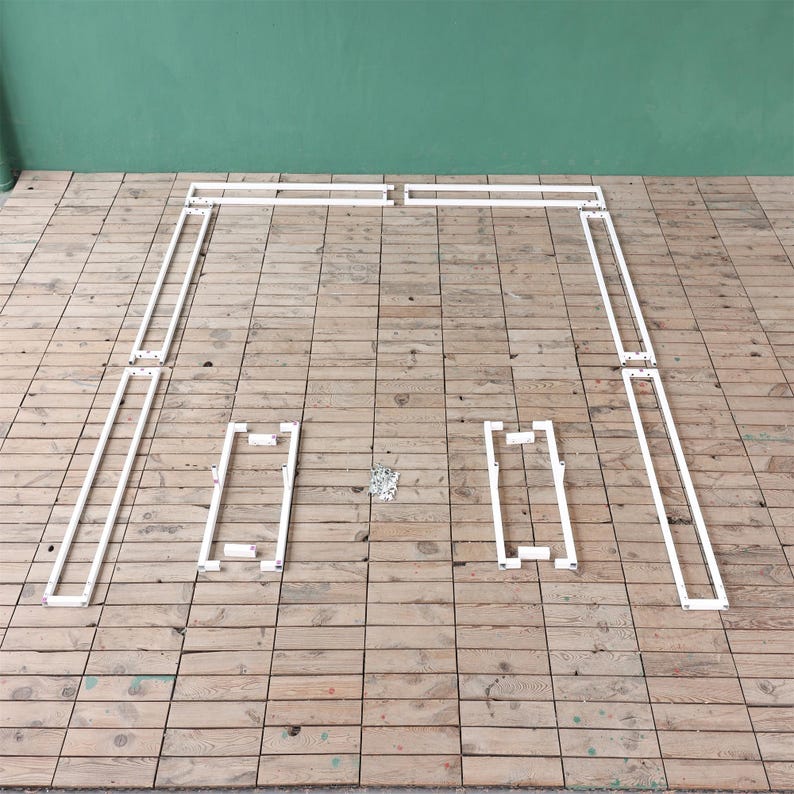 May include: White metal frame pieces laid out on a wooden floor. The pieces are in various sizes and shapes, and they appear to be part of a larger structure.