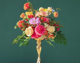 May include: A gold metal vase with a round top and three legs holds a bouquet of colorful silk flowers. The flowers include pink, red, yellow, orange, and white blooms.