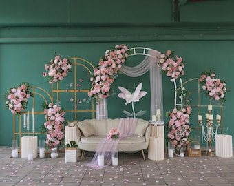 Puede incluir: Un telón de fondo para bodas o eventos con un sofá beige, arreglos florales y elementos decorativos. Rosas rosas adornan arcos dorados y blancos, con velas y una gran flor de papel. El suelo está cubierto de pétalos de rosa esparcidos.