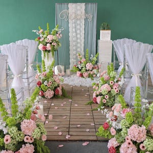 May include: A wedding ceremony setup featuring a wooden floor aisle lined with floral arrangements of pink hydrangeas, green bells of Ireland, and roses. White tulle-draped chairs flank the aisle, leading to a backdrop with a lace runner.