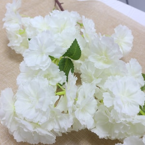 Pu&ograve; includere: Un mazzo di fiori artificiali bianchi con foglie verdi. I fiori sono raggruppati e hanno un aspetto delicato e stratificato.