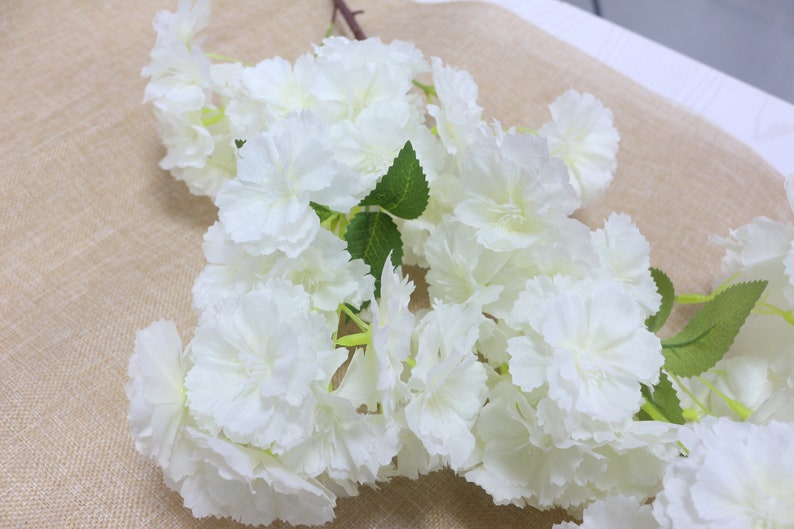 Pu&ograve; includere: Un mazzo di fiori artificiali bianchi con foglie verdi. I fiori sono raggruppati e hanno un aspetto delicato e stratificato.