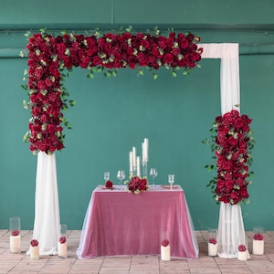 May include: A wedding arch adorned with deep red roses and foliage. The arch is draped with white fabric, set against a teal wall. A table is set with candles, glasses, and a floral centrepiece.