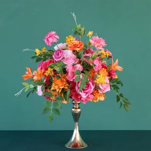 Könnte beinhalten: Ein großer Strauß aus rosa, orangefarbenen und gelben Blumen, die in einer goldenen Metallvase arrangiert sind. Die Blumen haben verschiedene Formen und Größen, und die Vase ist hoch und schlank.