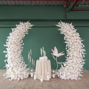 Könnte beinhalten: Ein Hochzeitszeremonien-Hintergrund mit zwei weißen Blumenbögen, einem weißen Tisch mit einer Spitzentischdecke und einer rosafarbenen Schmetterlingsdekoration.