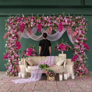 May include: A floral archway and backdrop with pink and magenta flowers, sheer white fabric, and a beige sofa. The scene is decorated with candles, flower arrangements, and scattered rose petals, creating a romantic setting.