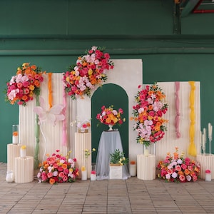 May include: A colourful floral display featuring bouquets of pink, orange, and yellow flowers. The display includes decorative panels, pedestals, and candles, creating a festive atmosphere. The scene is set against a green backdrop.