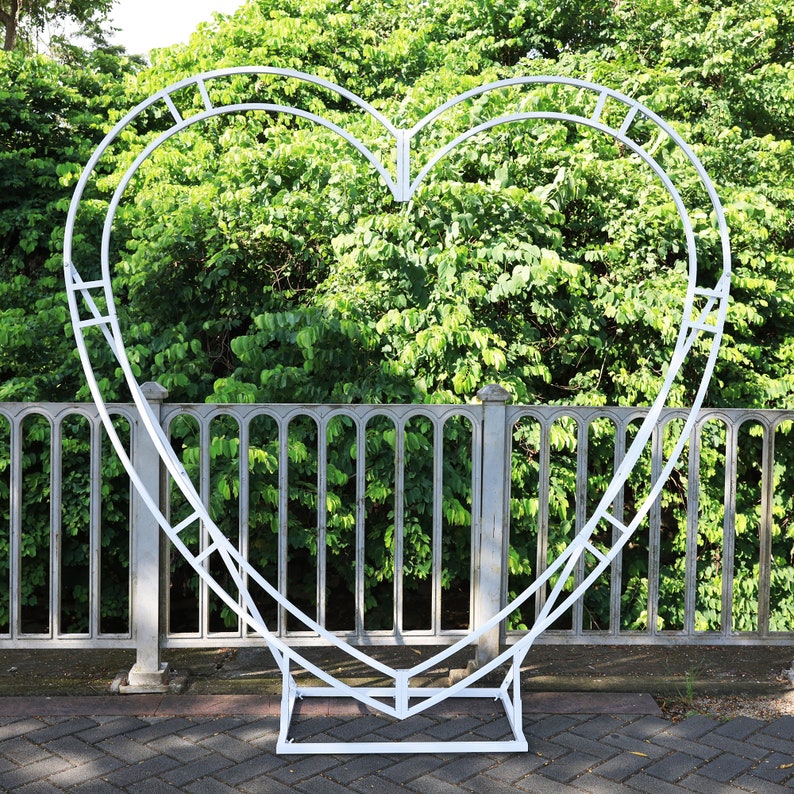 May include: White metal heart-shaped archway, perfect for a wedding or other special event. The archway is freestanding and has a simple, elegant design.