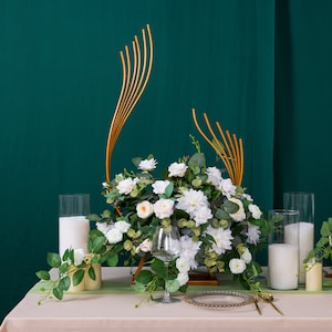 Könnte beinhalten: Ein goldfarbener Metallständer mit einem Blumenarrangement aus weißen und grünen Blumen. Der Ständer steht auf einem Tisch mit einer weißen Tischdecke. Auf dem Tisch stehen zwei klare Glaskerzenhalter mit weißen Kerzen.