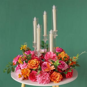 Peut inclure: Un bougeoir en verre transparent avec six bougies blanches entourées d'un arrangement floral de fleurs roses, orange et jaunes. Les fleurs sont disposées en cercle sur une table blanche.