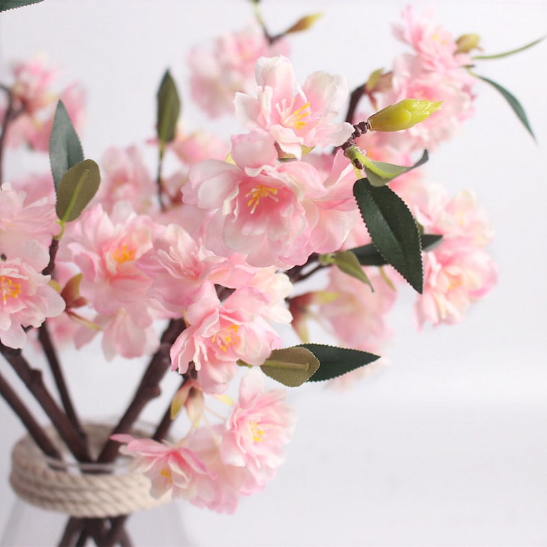 May include: A bouquet of pink artificial cherry blossoms with green leaves. The flowers are arranged in a vase with a rope trim.