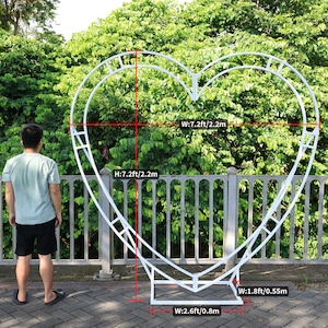 May include: White metal heart-shaped archway with measurements in feet and inches. The archway is 7.2 feet tall and 7.2 feet wide. The base of the archway is 2.6 feet wide and 1.8 feet deep.
