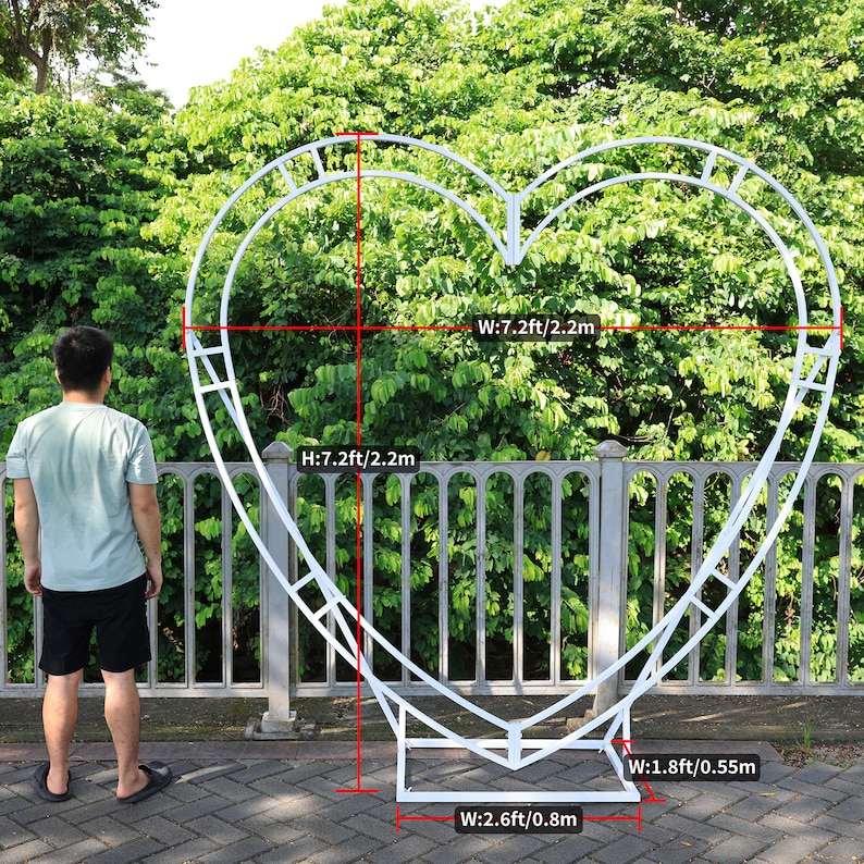 May include: White metal heart-shaped archway with measurements in feet and inches. The archway is 7.2 feet tall and 7.2 feet wide. The base of the archway is 2.6 feet wide and 1.8 feet deep.