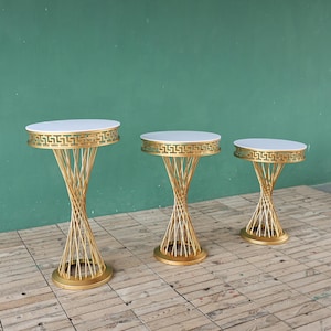 Peut inclure: Trois tables d'appoint en métal doré avec des plateaux blancs. Les tables ont une base en spirale et un motif grec décoratif sur le bord supérieur. Les tables sont de tailles différentes et reposent sur une surface en bois.