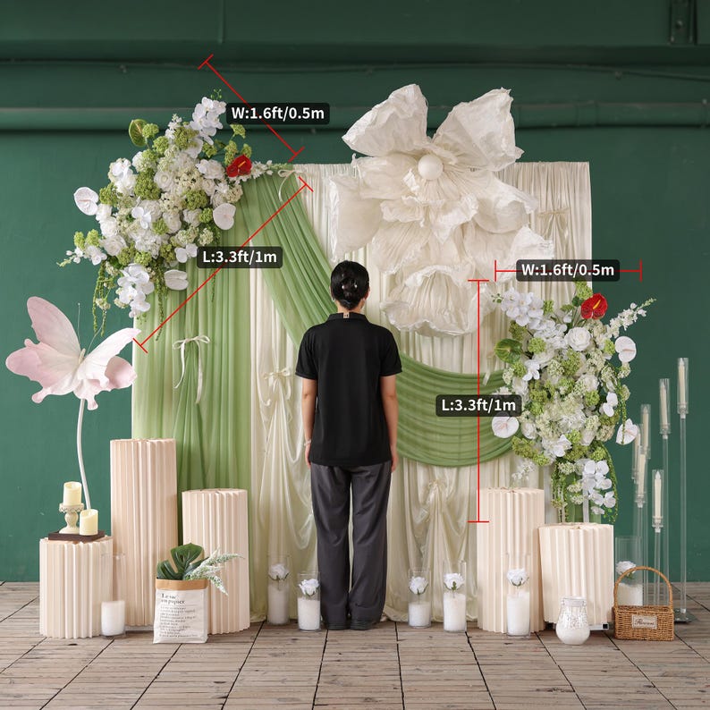 May include: A decorative backdrop for events, featuring floral arrangements, a large paper flower, and pleated pedestals. The backdrop includes green and white draping, with dimensions of 3.3ft/1m in length and 1.6ft/0.5m in width.