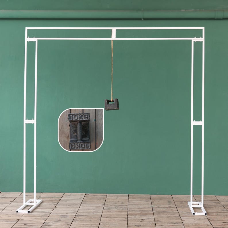 May include: White metal archway backdrop stand with two adjustable legs. A string hangs from the top of the archway with a weight attached to the end. The weight is a black metal block with the text "10KG" printed on it.