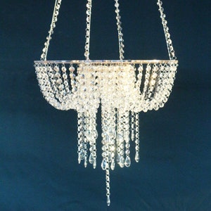 Puede incluir: Una lámpara de araña de cristal con un marco plateado y múltiples hebras de cuentas de cristal que cuelgan. La lámpara de araña está suspendida del techo por tres cadenas.
