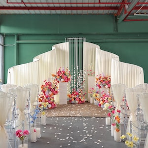 May include: A wedding ceremony setup with a backdrop of flowing white fabric and a stage adorned with colorful floral arrangements. Clear chairs are arranged in rows, and the floor is scattered with petals.