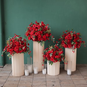 Könnte beinhalten: Arrangement aus roten Rosen auf cremefarbenen, geriffelten Sockeln. Klare Glaszylinder mit weißen Kerzen sind dazwischen angeordnet. Die Blumendekorationen stehen vor einer dunkelgrünen Wand und einem Holzboden.