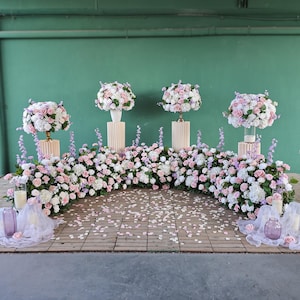 Puede incluir: Un arreglo floral con flores blancas y rosas, incluyendo rosas y hortensias, se exhibe sobre pedestales y a lo largo de la base. El fondo es una pared verde, y el suelo está cubierto de pétalos. También hay jarrones y velas.
