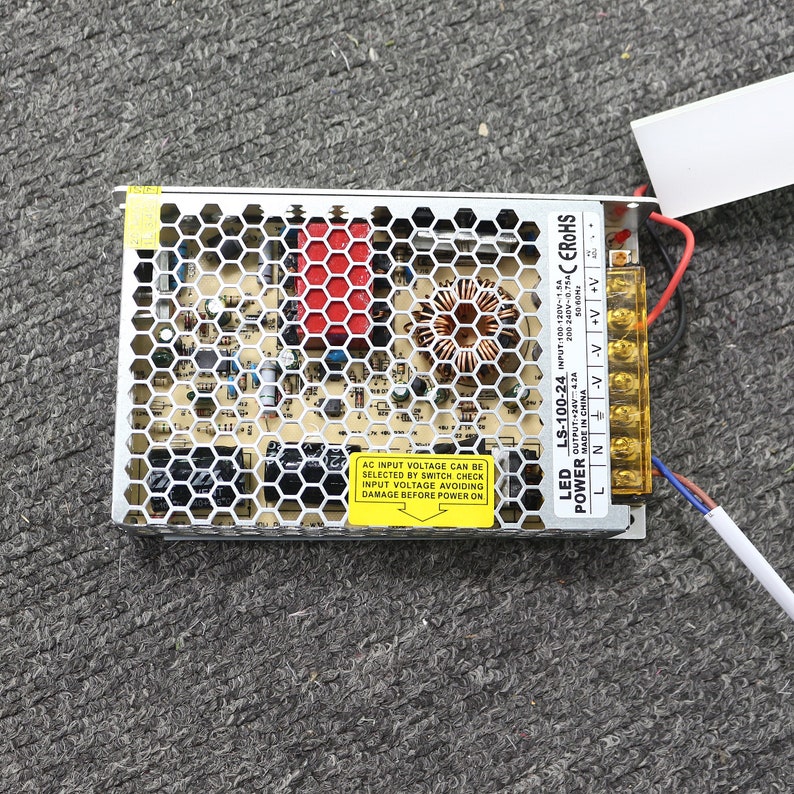 K&ouml;nnte beinhalten: Ein silbernes und schwarzes LED-Netzteil mit einem gelben Warnhinweis, der "AC input voltage can be selected by switch check input voltage avoiding damage before power on" lautet. Das Etikett hat einen roten Pfeil, der nach unten zeigt. Das Netzteil hat ein schwarzes Kabel und ein blaues und rotes Kabel, die daran angeschlossen sind. Das Netzteil tr&auml;gt den Text "LED LS-100-24 INPUT 100-120V-1.5A OUTPUT:+24V 4.2A 200-240V-0.75AC EROHS MADE IN CHINA", der darauf gedruckt ist.