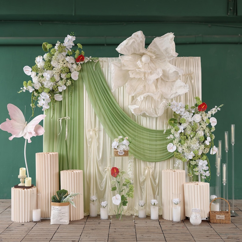 May include: A wedding backdrop featuring a cream-colored curtain with a green drape, adorned with white flowers, a large paper flower, and a pink butterfly. Beige pedestals and candles are arranged in front.