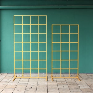 Puede incluir: Dos paneles de rejilla de metal dorado, uno más alto que el otro, de pie sobre un suelo de madera. Los paneles están hechos de finas barras de metal que crean un patrón de rejilla.