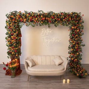 Puede incluir: Un sofá blanco con un arco floral detrás. El arco está hecho de vegetación y flores naranjas y rojas. El texto "Better Together" está escrito en letras de neón blancas en la pared detrás del sofá.